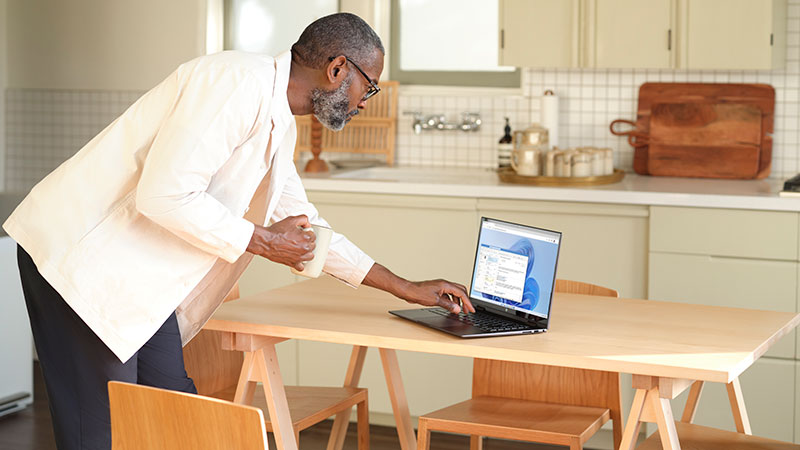 In a brightly lit modern kitchen, a man in a creme-colored blazer holds a coffee cup in one hand and types on a laptop PC sitting on a table.