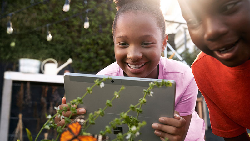 Two kids in a garden smile and use their Microsoft tablet to photograph a butterfly.