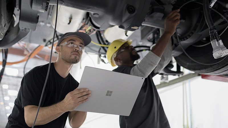 A man uses a Microsoft tablet while inspecting a vehicle alongside his coworker.