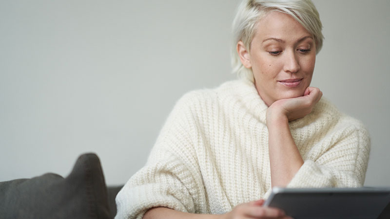 A woman inside sits on a couch and reads from a tablet.