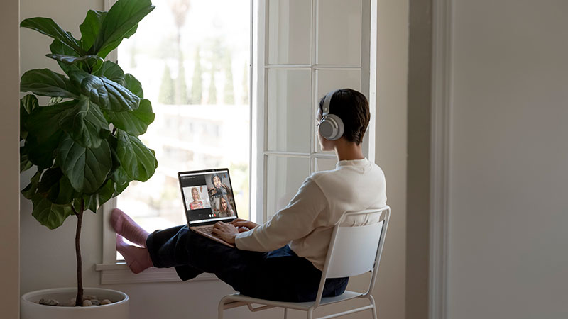 A woman seen from the back sits on a chair in front of a window while attending an online meeting.